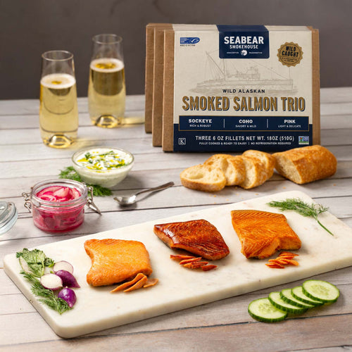 SeaBear Smokehouse Wild Alaskan Smoked Salmon Trio box featuring Sockeye, Coho, and Pink salmon fillets displayed on a marble serving platter alongside sliced baguette, dip, pickled red onions, and champagne glasses