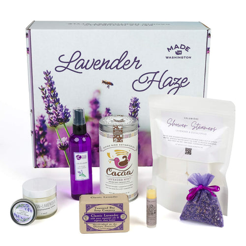 Lavender Haze gift set items displayed around the opened branded box, including room spray, shower steamers, lavender mint cacao mix, bar soap, lip balm, lotion, cuticle cream, and a purple sachet bag.