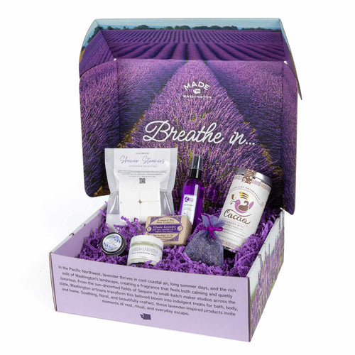 Lavender Haze gift set items displayed around the opened branded box, including room spray, shower steamers, lavender mint cacao mix, bar soap, lip balm, lotion, cuticle cream, and a purple sachet bag.
