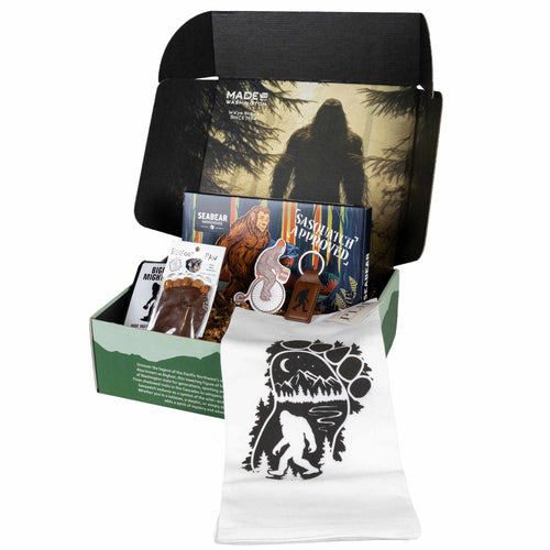 Legend of Sasquatch gift set items displayed around a light green "Made in Washington" box, including SeaBear smoked salmon, Bigfoot's Mighty Brew coffee, a Bigfoot Paw chocolate, a Sasquatch kitchen towel, a leather keychain, and a sticker.