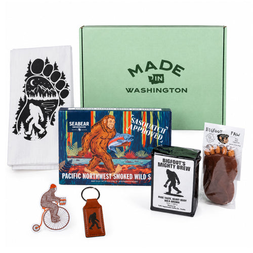 Legend of Sasquatch gift set items displayed around a light green "Made in Washington" box, including SeaBear smoked salmon, Bigfoot's Mighty Brew coffee, a Bigfoot Paw chocolate, a Sasquatch kitchen towel, a leather keychain, and a sticker.