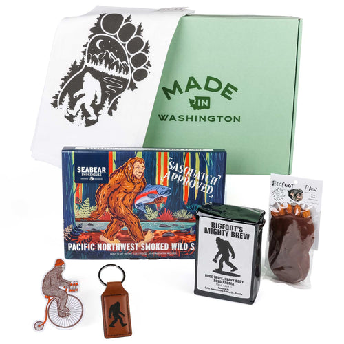 Legend of Sasquatch gift set items displayed around a light green "Made in Washington" box, including SeaBear smoked salmon, Bigfoot's Mighty Brew coffee, a Bigfoot Paw chocolate, a Sasquatch kitchen towel, a leather keychain, and a sticker.