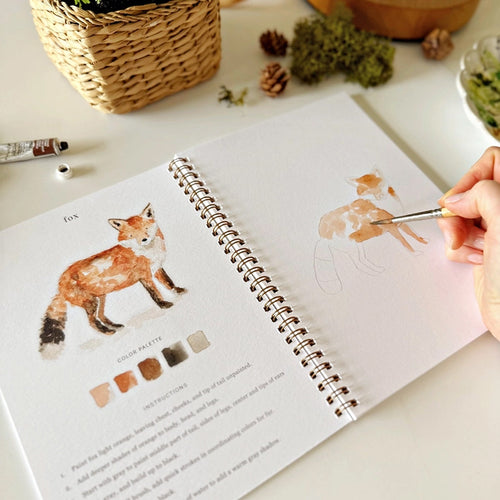 Notebook titled 'Woodland Watercolor Workbook' with watercolor swatches and a bear illustration, surrounded by nature-themed items. | Emily Lex Studios Woodland Watercolor Workbook | Made In Washington | PNW Gifts