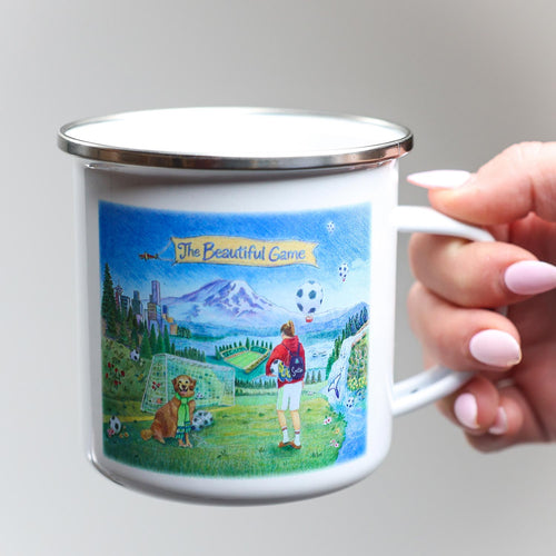 Enamel mug with 'The Beautiful Game' design featuring a soccer player and dog on a scenic background.