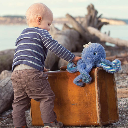 A light blue fluffy octopus plush toy with blue eyes, a small white paper tag attached, and blue-and-white striped accents on its arms, photographed on a plain white background.