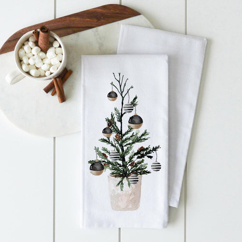 A white cotton kitchen towel with a watercolor illustration of a small Christmas tree in a pot with ornaments.