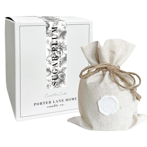 A lit white candle in a clear glass jar with the text 'SUGAR PLUM' and 'PLUM + BLACK FIG' printed on the front, shown against a plain white background.
