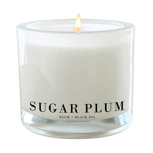A lit white candle in a clear glass jar with the text 'SUGAR PLUM' and 'PLUM + BLACK FIG' printed on the front, shown against a plain white background.