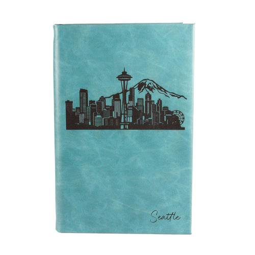 A topaz faux-leather notebook featuring a black silhouette of the Seattle skyline, including the Space Needle and Mount Rainier, with the word 'Seattle' written in black script near the bottom right.