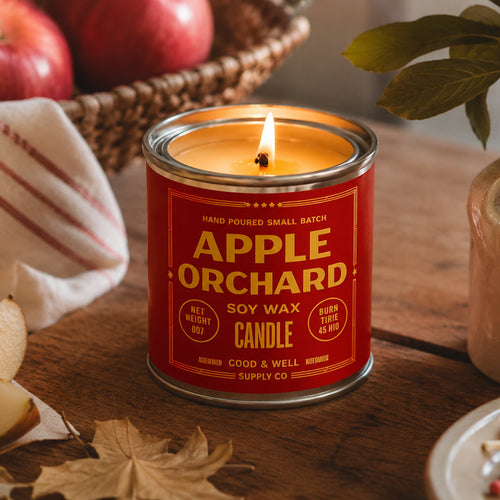 A small cylindrical candle tin with a bright red label and silver metal rim, reading 'APPLE ORCHID SOY WAX CANDLE' in yellow text, centered on a plain white background.