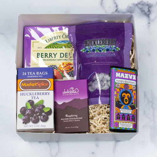 A curated berry-themed gift basket featuring a box of Liberty Orchards Berry Delight, a bag of Huckleberry Candy, a jar of Marionberry Jam , a box of Dolcetta Raspberry Chocolate bar, and a colorful Maeve Hungry Blackberry chocolate bar, arranged in front of a white 'Made Washington' box on a white background.