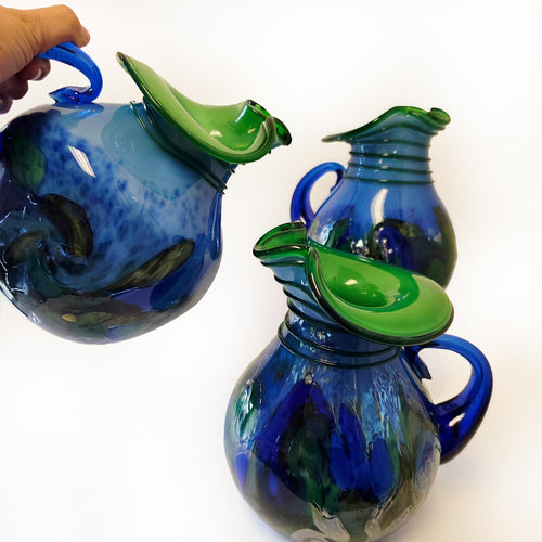 A decorative glass pitcher with a rounded body, a curved handle, and a wide flared spout lid, featuring an organic color pattern that transitions between deep cobalt blue and emerald green on a white background.