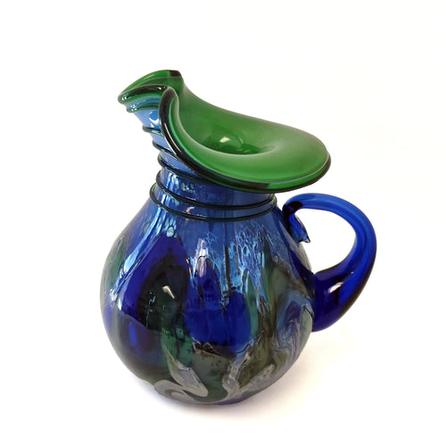A decorative glass pitcher with a rounded body, a curved handle, and a wide flared spout lid, featuring an organic color pattern that transitions between deep cobalt blue and emerald green on a white background.