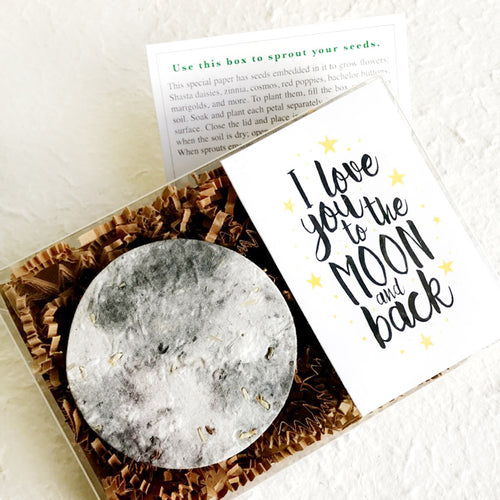 A plantable gift card featuring a large textured moon embedded with seeds on brown crinkle paper beside the phrase 'I love you to the MOON and back' in black script with small yellow stars on a white background.
