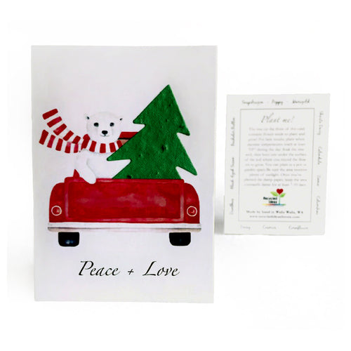 A Christmas greeting card featuring a white polar bear in a red pickup truck carrying a green plantable seed paper Christmas tree, with a red and white striped scarf and the words 'Peace + Love' printed below. A small printed back card is shown beside it on a white background.