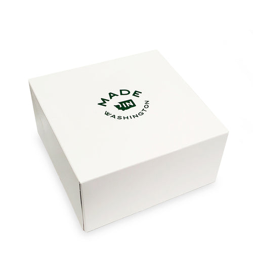 A curated wellness gift set featuring a white 'Made in Washington' box, a Rosemary-Mint moisturizing lotion, a Solstice bath salt jar, a Sugar Scrub jar, a wrapped Estrella bar soap, and a Santal & Rum candle, arranged on a white background.