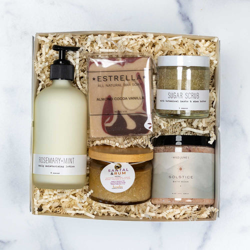 A curated wellness gift set featuring a white 'Made in Washington' box, a Rosemary-Mint moisturizing lotion, a Solstice bath salt jar, a Sugar Scrub jar, a wrapped Estrella bar soap, and a Santal & Rum candle, arranged on a white background.
