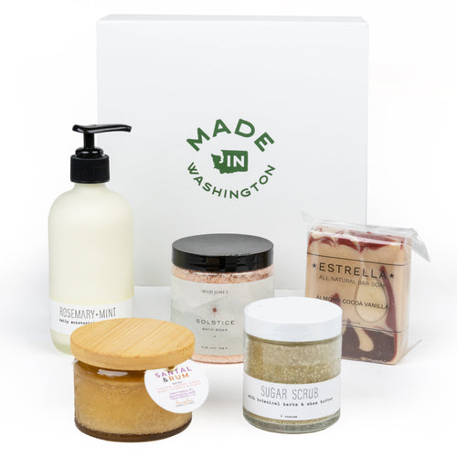 A curated wellness gift set featuring a white 'Made in Washington' box, a Rosemary-Mint moisturizing lotion, a Solstice bath salt jar, a Sugar Scrub jar, a wrapped Estrella bar soap, and a Santal & Rum candle, arranged on a white background.