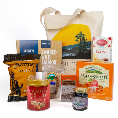 A curated gift collection of iconic Washington brands featuring a large beige canvas tote bag with a yellow and green coastal landscape print, surrounded by assorted snack and food items including a bag of Harvey's popcorn, Seabear smoked wild salmon, Fisher scone, Almond Roca, and a jar of Huckleberry jam, all arranged on a white background.