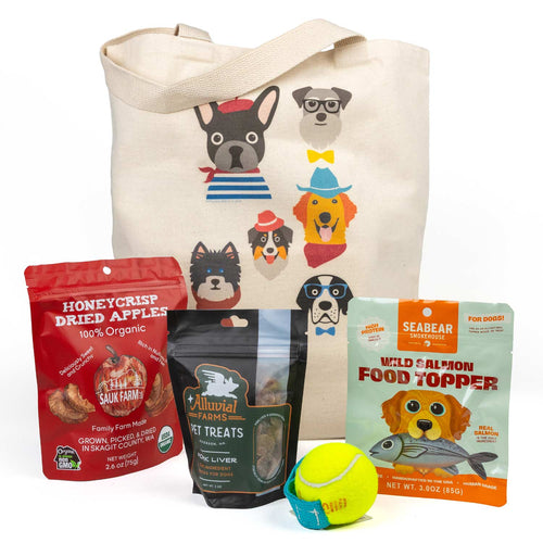 A dog-themed gift set featuring a beige canvas tote bag with colorful dog illustrations, a red bag of Honeycrisp Dried Apples, a black bag of Alluvial Pet Liver Treats, a Seabear Wild Salmon Food Topper bag, and a yellow tennis ball fetch toy.