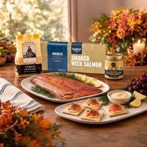 A gourmet food gift set arranged on a wooden table with seasonal decor. Visible products include a bag of Beecher's Original Crackers, a SeaBear Smoked Wild Salmon box, and a jar of AJ's Walla Walla mustard. alongside plated smoked salmon, crackers, lemon wedges, and a small bowl of mustard.