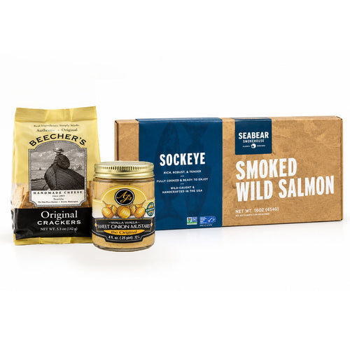 A gourmet food gift set arranged on a wooden table with seasonal decor. Visible products include a bag of Beecher's Original Crackers, a SeaBear Smoked Wild Salmon box, and a jar of AJ's Walla Walla mustard. alongside plated smoked salmon, crackers, lemon wedges, and a small bowl of mustard.
