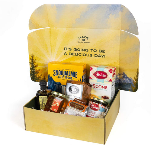 Assorted food and beverage products including scones, maple syrup, coffee, jam, and tea on a white background in front of a box designed with a watercolor-like Pacific Northwest forest and mountain scene and the words Breakfast In A Box.