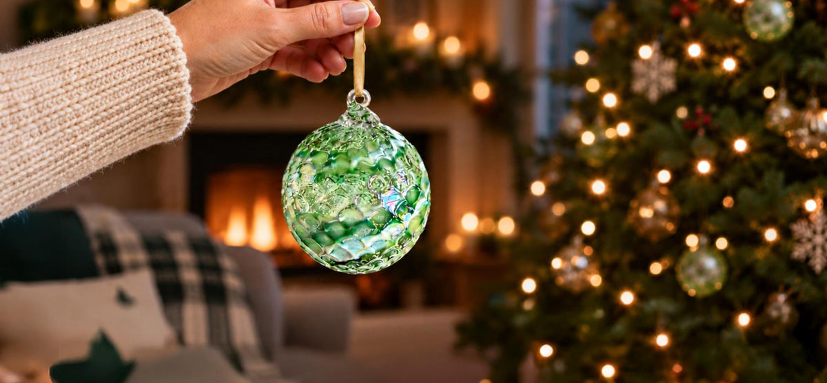 Shop the Glass Eye Studio 2026 Limited Edition green blown glass ornament held in front of a holiday tree.