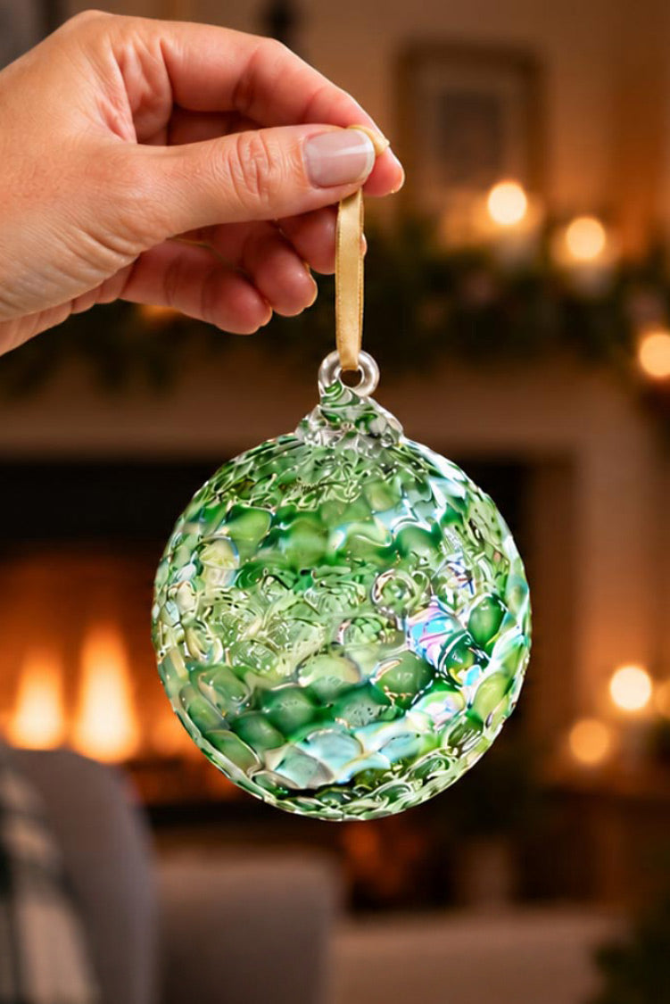 Shop the Glass Eye Studio 2026 Limited Edition green blown glass ornament held in front of a holiday tree.