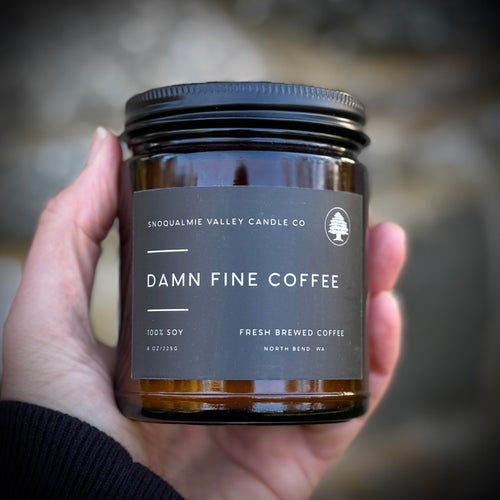 hand holding a brown glass jar candle with a black lid, the label reads Damn Fine Coffee by Snoqualmie Valley Candle Co.