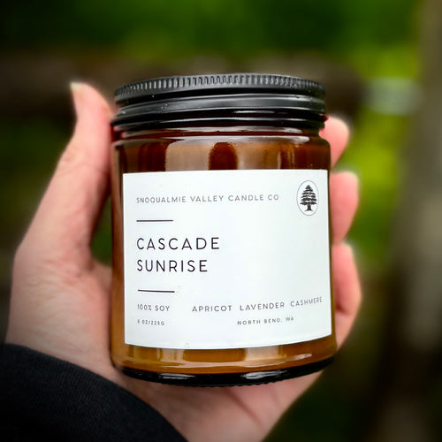 A hand holding a glass jar candle with a black metal lid and a white label that reads 'Cascade Sunrise' by Snoqualmie Valley Candle Co, against a blurred green outdoor background.