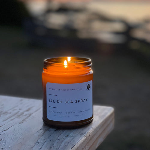 A lit amber glass jar candle labeled 'Salish Sea Spray' by Snoqualmie Valley Candle Co., sitting on a weathered wooden surface with a blurred outdoor background.