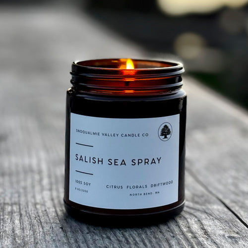 A lit amber glass jar candle labeled 'Salish Sea Spray' by Snoqualmie Valley Candle Co., sitting on a weathered wooden surface with a blurred outdoor background.