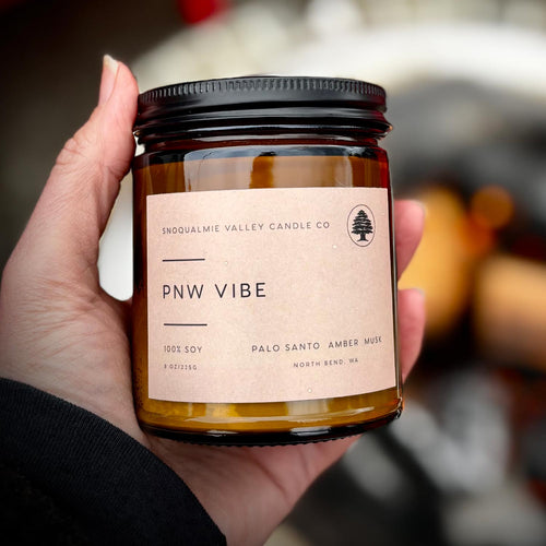 A hand holding a soy candle in an amber glass jar with a black lid and a beige paper label that reads 'SNQUALMIE VALLEY CANDLE CO. PNW VIBE'.