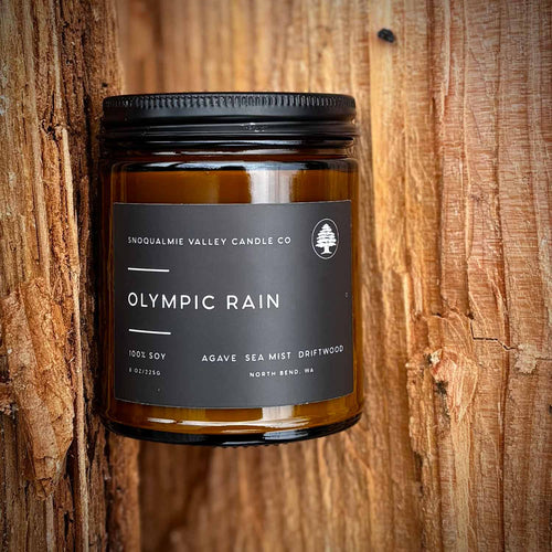 An amber glass jar candle with a black metal lid and a dark charcoal label reading 'Olympic Rain' by Snoqualmie Valley Candle Co, photographed against a rustic wood plank background.