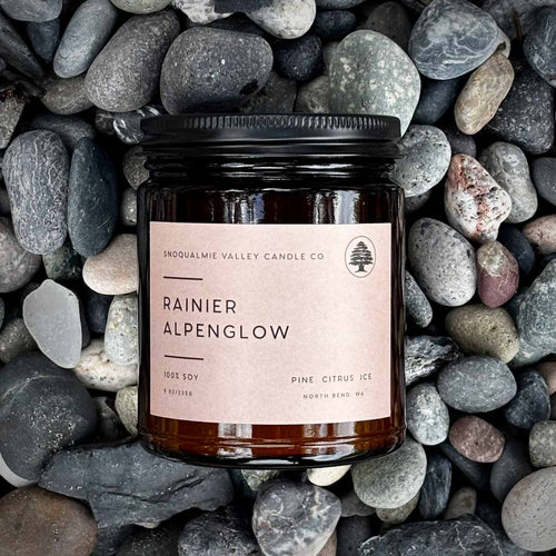 A soy candle in a dark amber glass jar with a black lid and a beige label reading 'Rainier Alpenglow' by Snoqualmie Valley Candle Co, photographed from above on a bed of smooth gray and black stones.