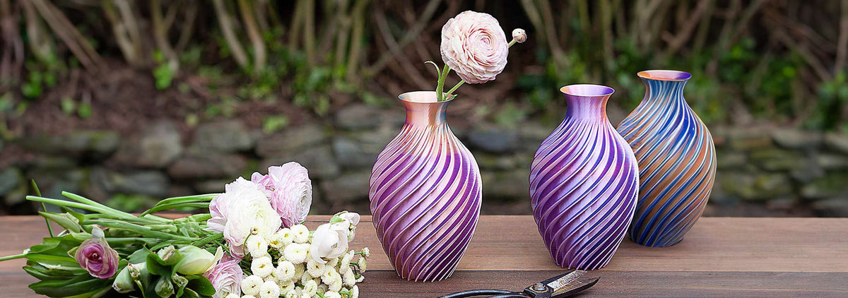 Three handcrafted spiral 3-D printed vases with iridescent purple and copper finishes on a table next to fresh flowers.
