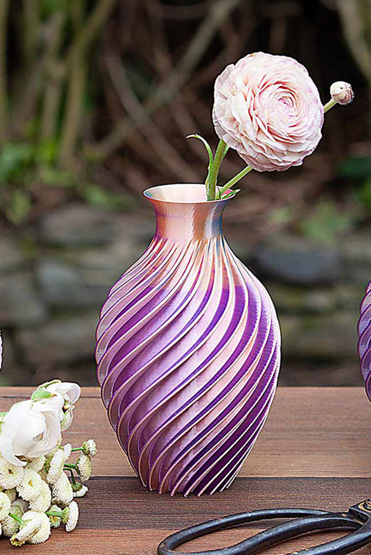 Three handcrafted spiral 3-D printed vases with iridescent purple and copper finishes on a table next to fresh flowers.