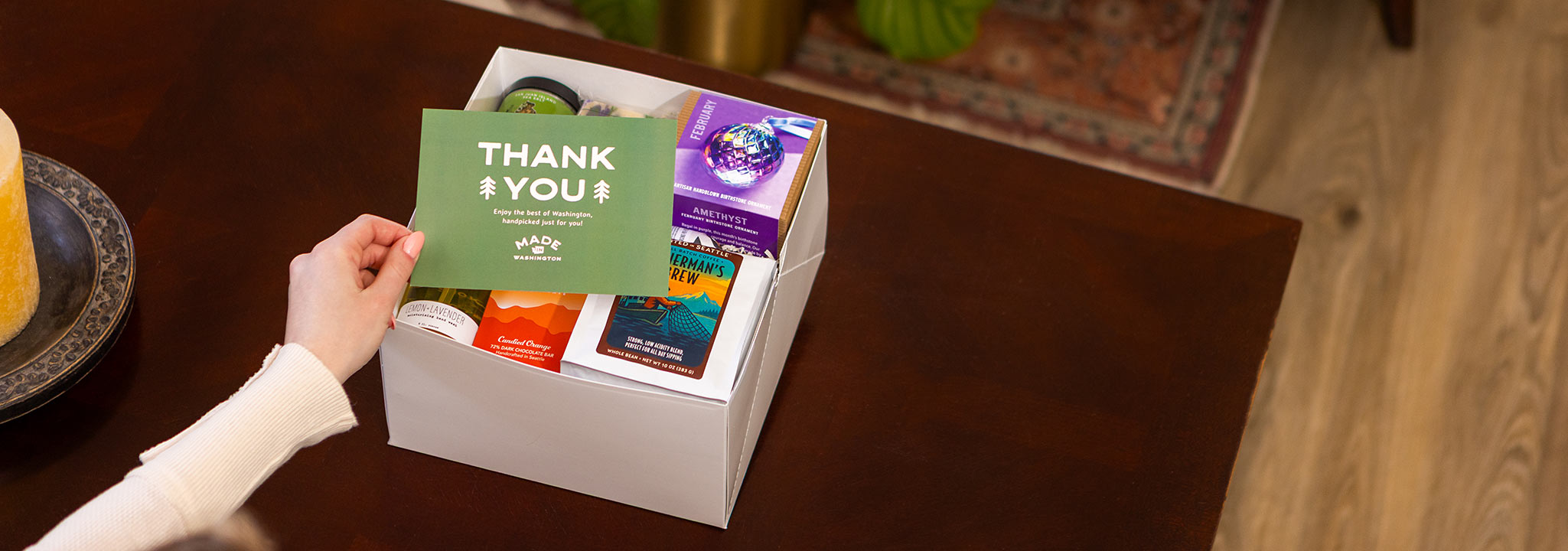 Person holding a 'Thank You' card over a gift box with snacks on a wooden table. | Buy Local Gifts At Made In Washington Gift Stores