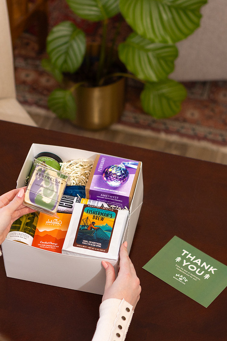 Person holding a box of filled with locally made foods & gifts with a 'Thank You' card on a wooden surface | Shop for locally made artisan gifts at Made In Washington Gift Stores.